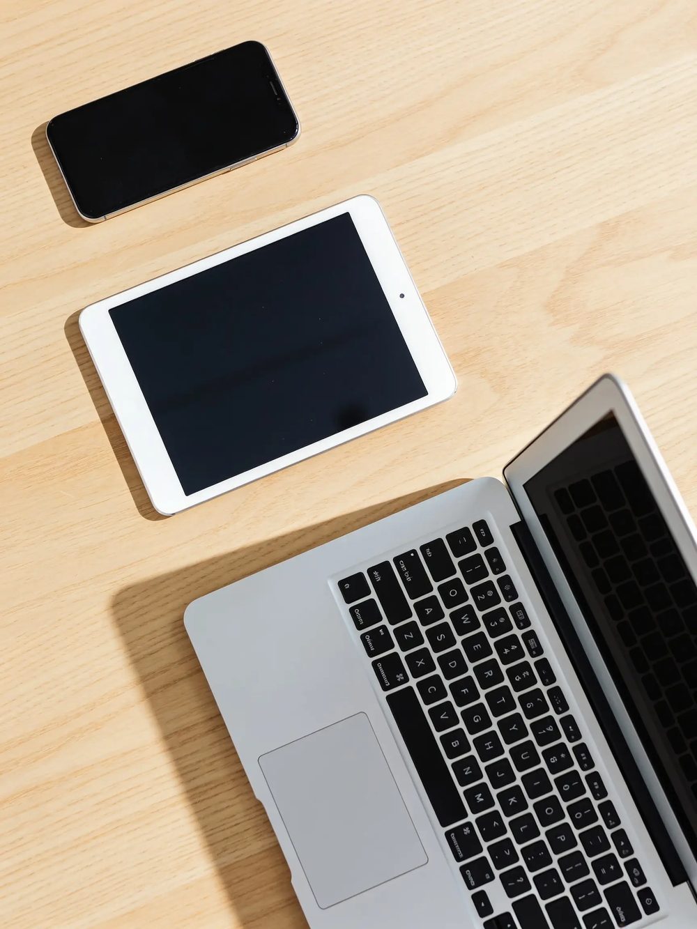 A phone, a tablet and a laptop laid out in a row on a wooden desk